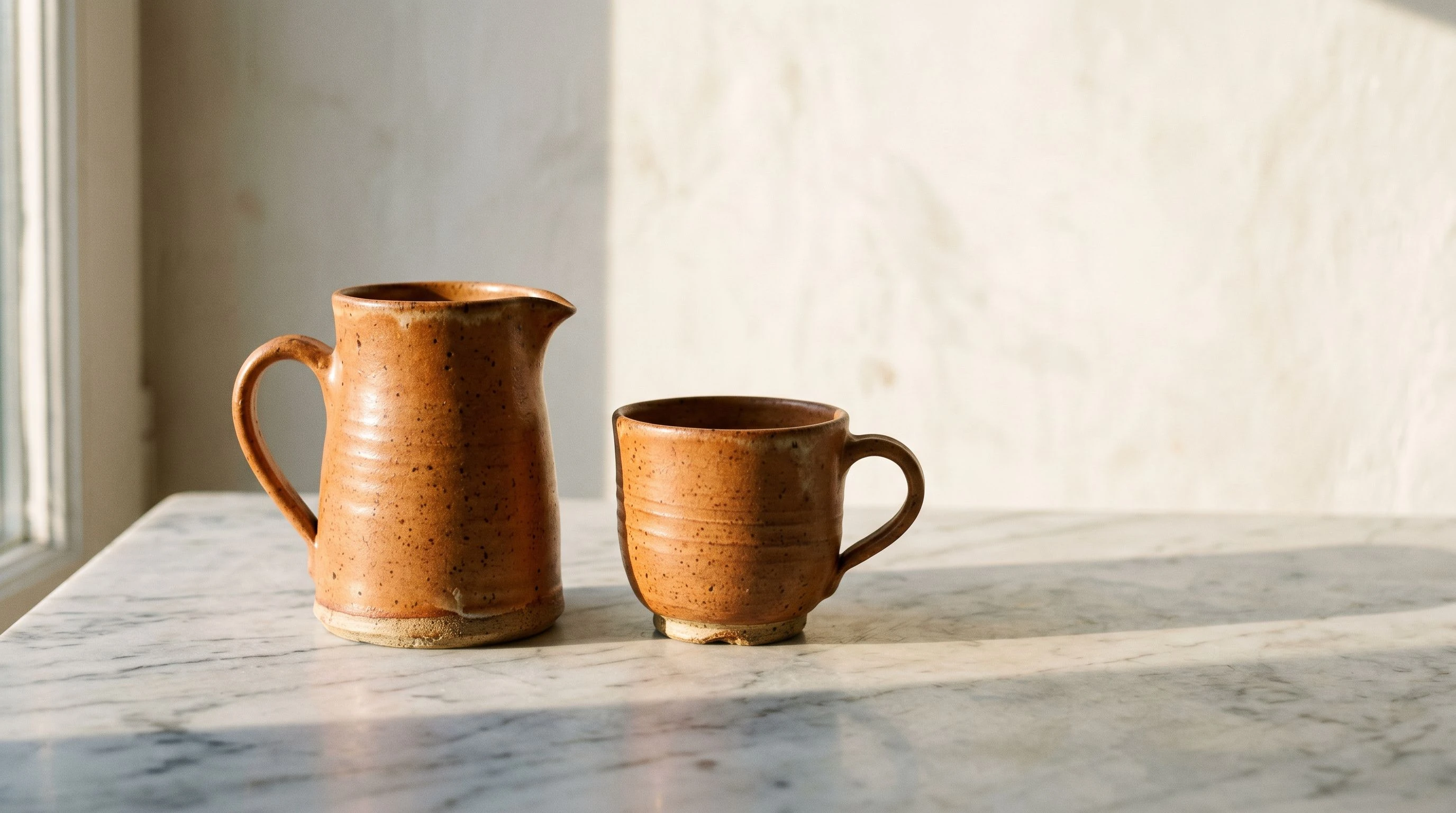 Two ceramic vessels — a pitcher and a cup — in terracotta glaze, styled together on a marble surface